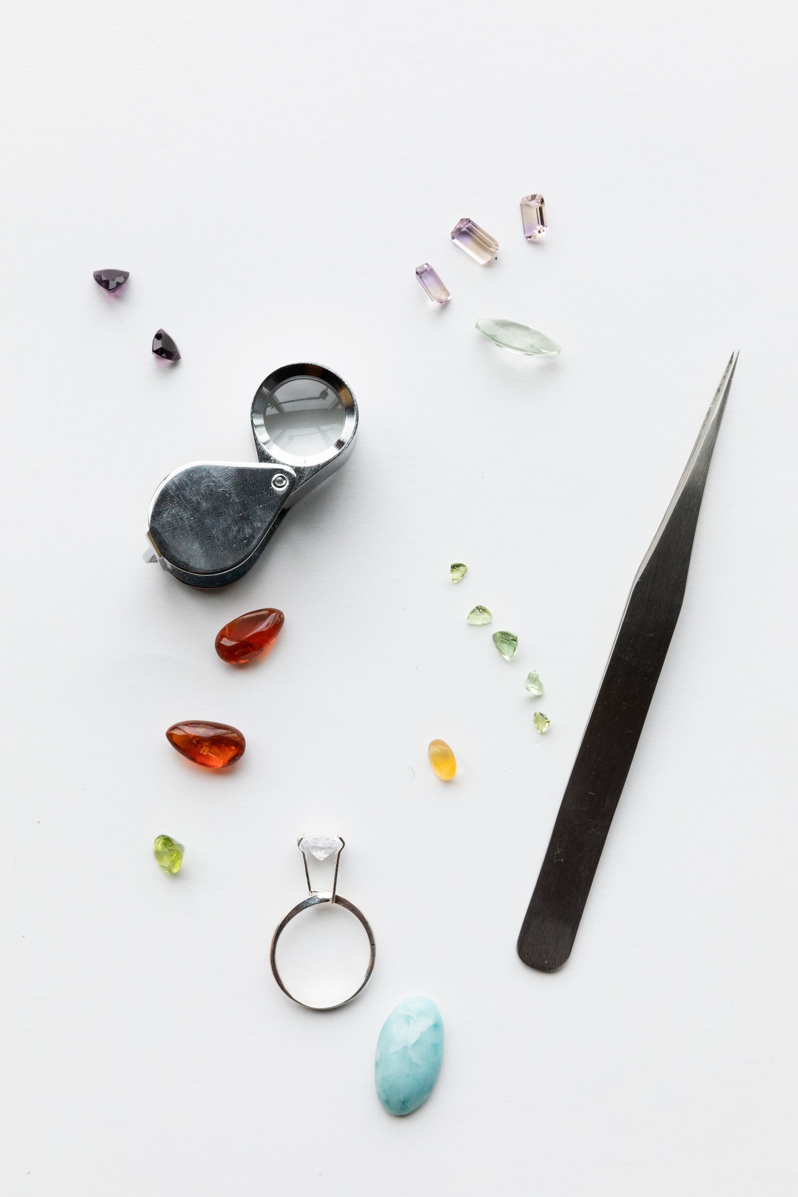 Loose coloured gemstones beside a jeweller's loupe, tweezers and a solitaire ring